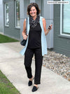 Woman standing in front of a building wearing Miik's Avy button-up dressy tunic top in blue as a cardigan with an all black outfit underneath.