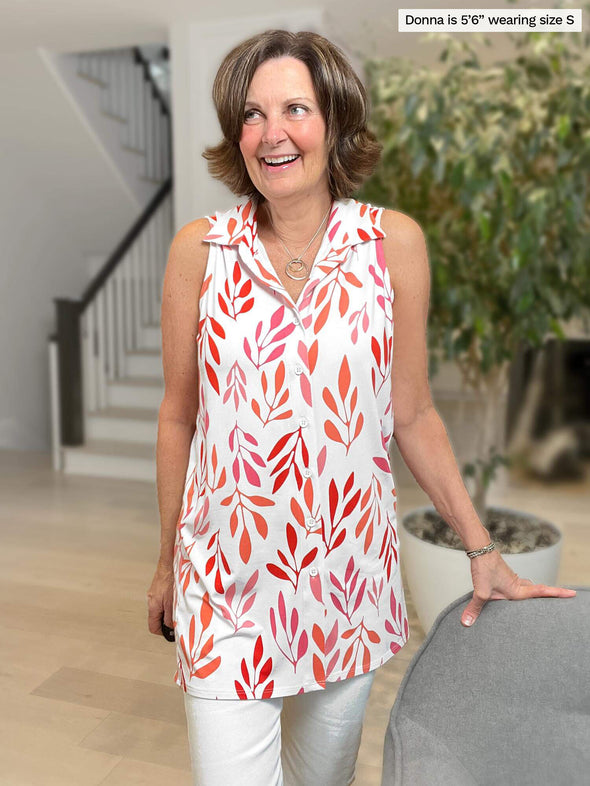 Woman standing in the living room looking away laughing while wearing Miik's Avy button-up dressy tunic top in pink leaf pattern and white pants.