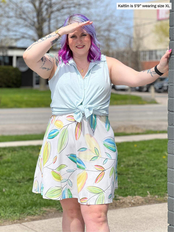 Woman standing next to a brick wall wearing Miik's Avy button-up dressy tunic top in light blue tied at the waist over a colour full leaf pattern dress.