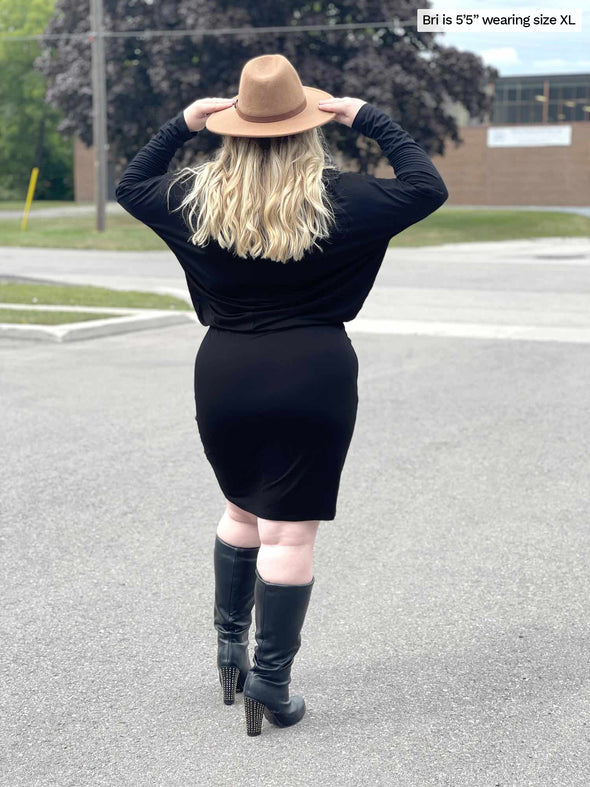 Woman standing backwards to the camera holding her hat showing the back of Miik's Bali bat sleeve dress in black and high heels boots