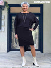 Woman smiling standing in front of a doorway wearing Miik's Bali batwing sleeve dress in black with high heels boots