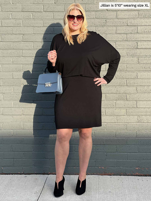 Woman standing in front of a brick wall wearing Miik's Bali batwing sleeve dress in black while holding a blue purse 