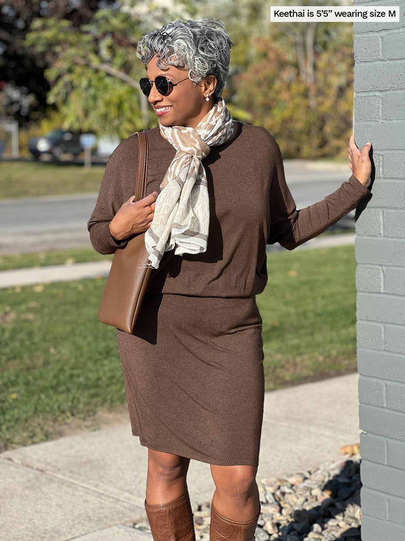 Woman standing next to a wall wearing Miik's Bali batwing sleeve dress in brown with a scarf around her neck.  