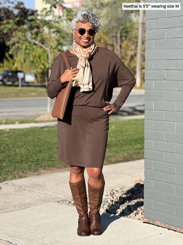 Woman standing next to a wall wearing Miik's Bali batwing sleeve dress in brown with a scarf around her neck.