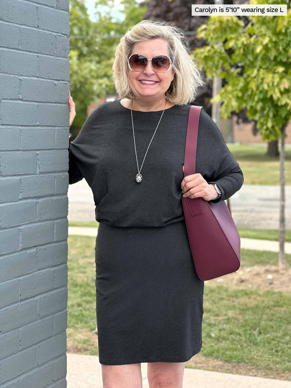 Woman standing next to a brick wall wearing Miik's Bali bat sleeve dress in charcoal with a port shoulder bag and sunglasses 