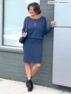 Woman standing next to a wall wearing Miik's Bali batwing sleeve dress in navy melange.