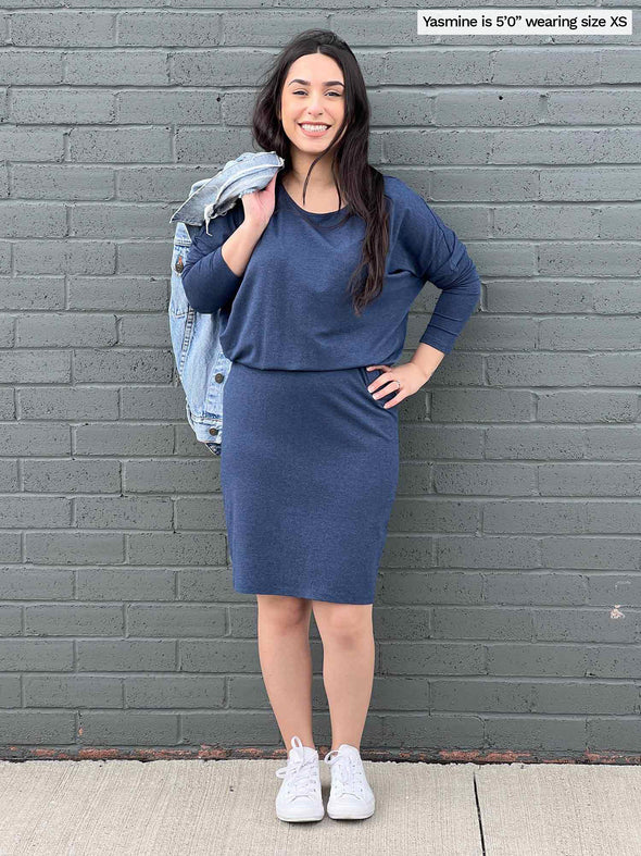 Woman standing in front of a wall wearing Miik's Bali batwing sleeve dress in navy melange with a jean jacket thrown over her shoulder.