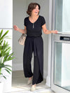 Woman standing at the doorway smiling and wearing Miik's black blair tie belt and black Marianna reversible classic tee with black Shea Tulip Pants while smiling and holding her purse