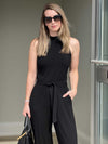 Woman standing in front of a beige wall wearing Miik's black blair soft tie belt with the black samra mockneck sleeveless top with black pants while holding a purse and wearing sunglasses