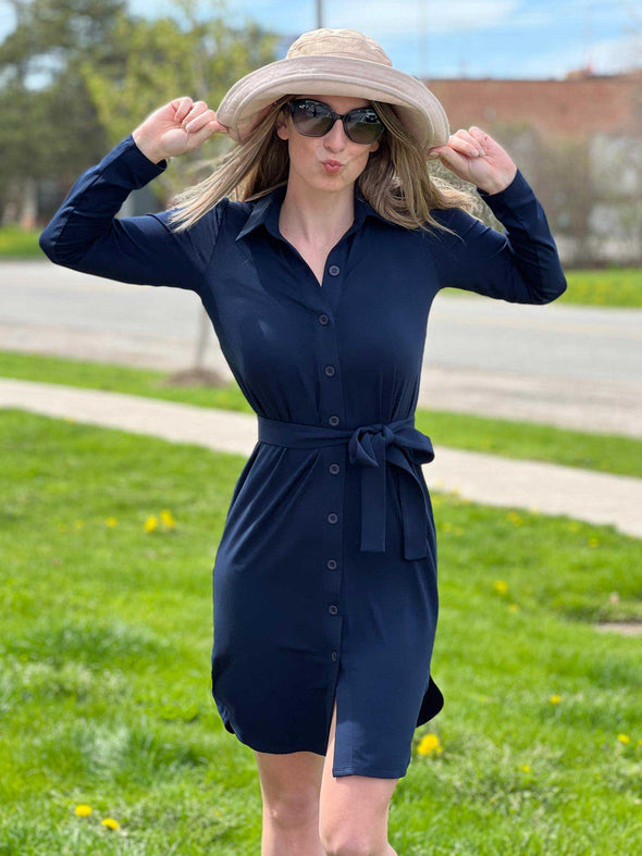 Woman standing outside holding her hat and wearing Miik's navy blair soft tie belt with the navy Flynn collared button-up shirt dress