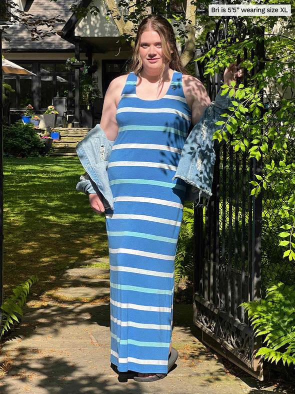 Woman standing in nature by a gate wearing Miik's brodie racerback maxi tank dress in blue stripe with a jean jacket