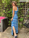 Woman smilling and standing sideways showing off the side slits in front of trellis wearing Miik's brodie racerback tank maxi dress in blue stripe color