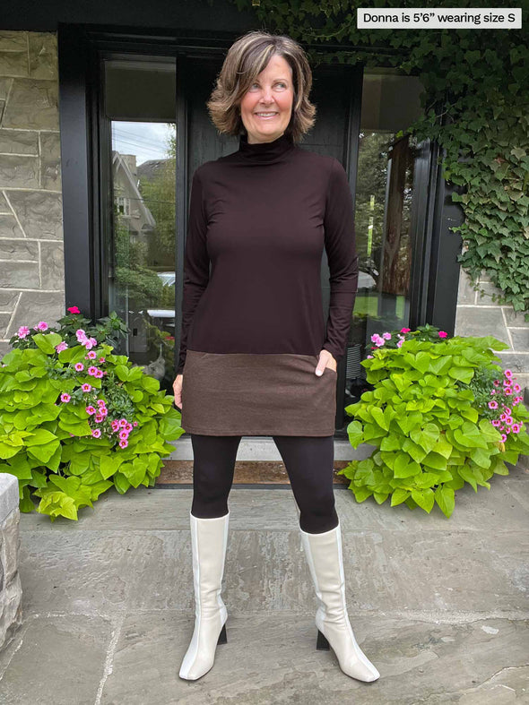 Woman standing in a backyard wearing Miik's Brooklin mock neck pocket tunic in dark chocolate/chocolate melange with leggings and beige boots
