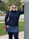 Woman standing next to a brick wall and smiling wearing Miik's Brooklin mock neck pocket tunic in navy/navy melange with a matching colour legging