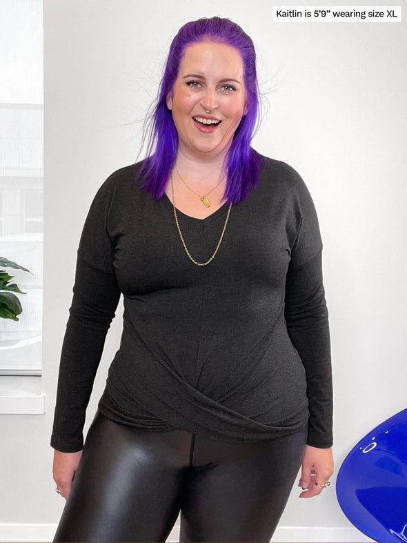 Woman standing in front of a wall wearing Miik's Chenley twist-front top in grey with leather leggings.