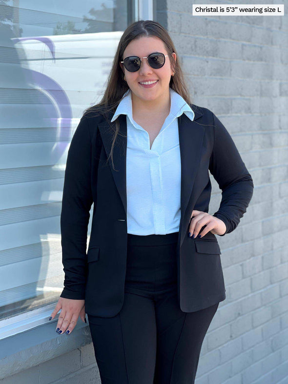 Woman standing next to a window smiling wearing a light blue collared shirt with Miik's Danica lightweight tailored blazer with notched collar in black with matching black pants.
