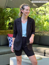 Woman standing in backyard wearing a light blue top tied with Miik's Danica lightweight tailored blazer with notched collar in black with black biker shorts.
