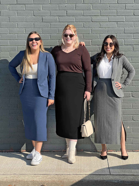 Three women smiling standing in front of a brick wall all wearing Miik's Devon midi skirt in three different collours: navy melange, black and granite pinstripe
