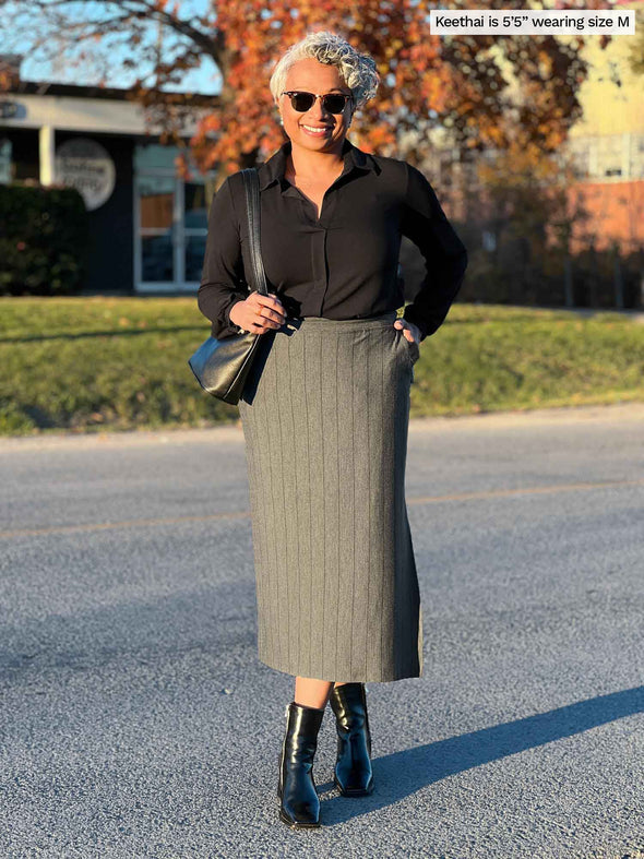 Woman smiling wearing Miik's Devon midi skirt in granite pinstripe with a collared black top and matching colour boots and purse.