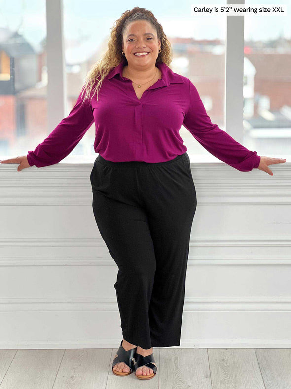 Miik model Carley (five feet two, size double extra large) smiling while standing with open arms in front of a window wearing Miik's Easton pocket capri in black and a collared shirt um ruby and sandals