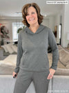 Miik founder Donna (5'6, small) smiling while leaning against to a couch wearing Miik's Essex luxe fleece hoodie in granite melange size medium for a more overzied look