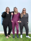 Three women standing in front of a grey wall wearing Miik's Fraya stretchy lounge jumpsuit in three different colours: black, port melange and charcoal