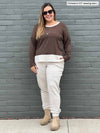 Woman standing in front of a brick wall wearing Miik's Geneva two-tone crew neck top in brown chocolate melange/oatmeal, oatmeal fleece jogger, boots and sunglasses 