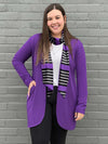 Woman standing in front of a brick wall smiling while wearing Miik's halona scarf in purple, along with a long purple cardigan