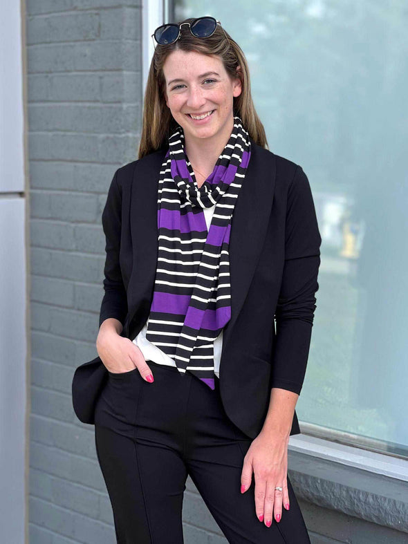 Woman standing in front of a window smiling while wearing Miik's Halona tie scarf in purple, along with a black blazer and a matching colour pants 