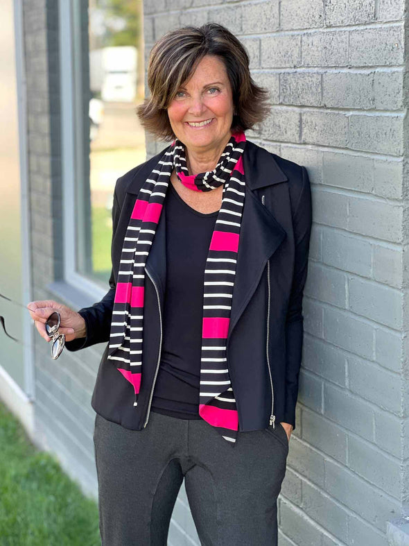 Woman smiling while leaning against a brick wall wearing Miik's Halona tie scarf in raspberry stripe, along with a jacket and top in black and charcoal pants  