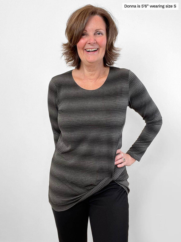 Woman standing in front of a wall wearing Miik's Harmony twisted hem top in grey stripe with black pants.