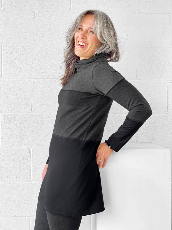 Woman standing in front of a wall wearing Miik's Hasha colourblock twisted cowl tunic in grey stripe and leggings.