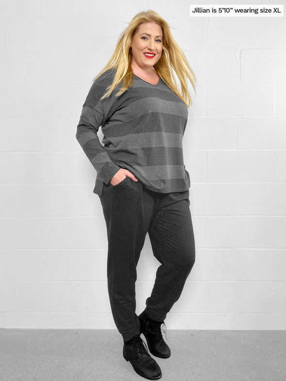 Woman standing sideway in front of a white wall wearing Miik's Henley lounge jogger in charcoal with a striped long sleeve v neck top