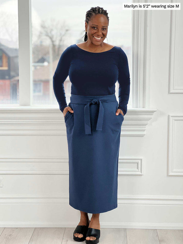 Woman smiling standing in front of a window/white wall wearing Miik's Hilden ballet top in navy along with a midi skirt in navy melange and hands on its pockets 
