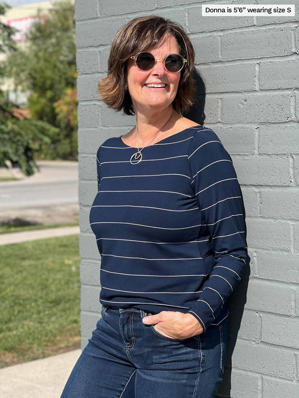 Woman smiling leaning against to a brick wall wearing Miik's Hilden ballet top in navy pinstripe, jeans and sunglasses
