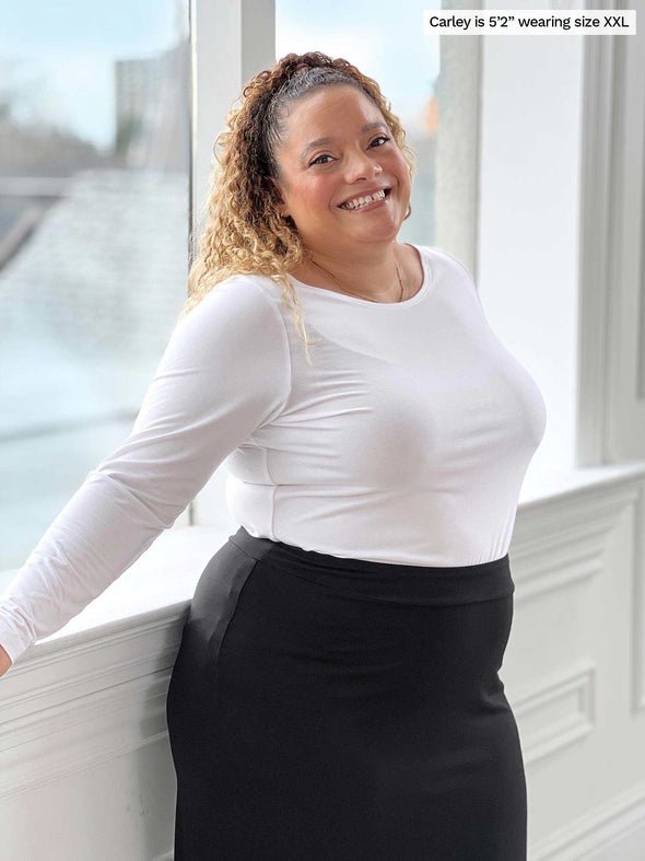 Miik model Carley (five feet two, size double extra large) smiling while standing in front of a window wearing Miik's Hilden ballet top in white tucked in a midi skirt in black