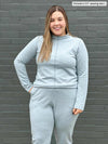 Woman standing in front of a brick wall wearing Miik's India reversible fleece funnel neck sweater in mist melange with a matching colour fleece jogger 