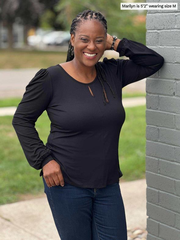 Woman smiling while leaning against a brick wall wearing Miik's Janette top in black and jeans