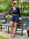 Woman leaning on the back of a garden chair with a pool in the background, smiling and looking away while wearing Miik's Janice 3/4 sleeve short romper in navy with a brown belt and sandals.