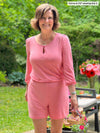 Woman standing in nature with chairs in the background smiling and wearing Miik's Janice 3/4 sleeve short romper in pink colour and holding her sandals.