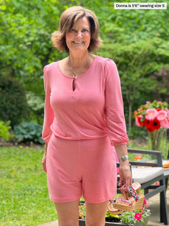 Woman standing in nature with chairs in the background smiling and wearing Miik's Janice 3/4 sleeve short romper in pink colour and holding her sandals.