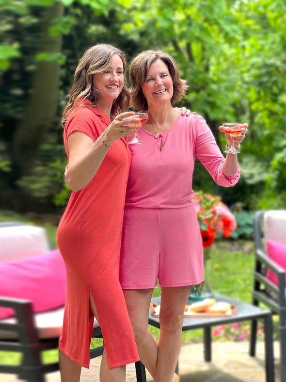 Two women standing in front of garden chairs looking away and raising their drinks while wearing Miik's Janice 3/4 sleeve short romper in pink, with the Dylan collared midi sheath dress in orange colour.