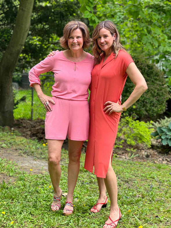 Two women standing in nature smiling and wearing Miik's Janice 3/4 sleeve short romper in pink, along with Dylan collared midi sheath dress in orange colour.