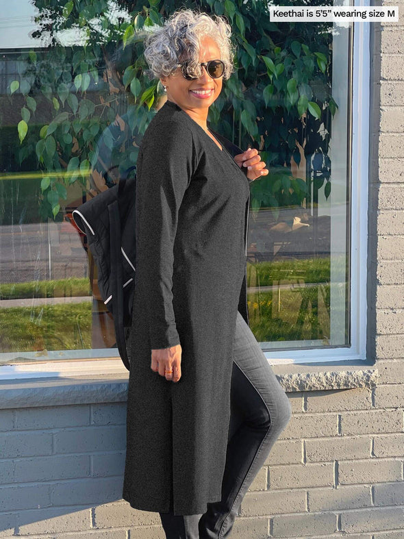 Woman standing sideway in front of a window wearing Miik's Joey long duster cardigan in charcoal and jeans