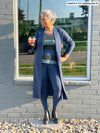 Woman standing in front of a window while holding a glass wearing Miik's Joey long duster cardigan in navy melange with a matching colour legging and a striped tank top