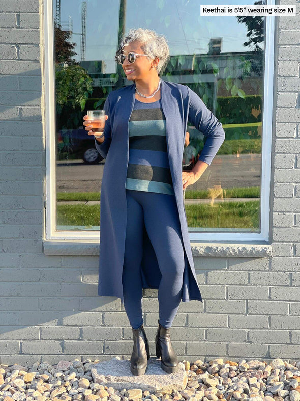 Woman standing in front of a window while holding a glass wearing Miik's Joey long duster cardigan in navy melange with a matching colour legging and a striped tank top