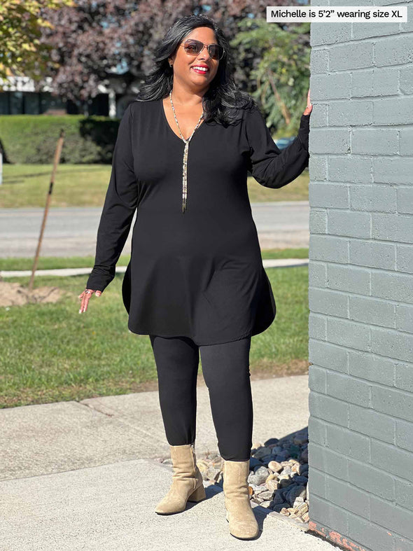 Woman standing next to a building wearing Miik's Juno reversible long sleeve tunic in black with black leggings. 