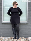 Woman standing in front of a window wearing Miik's Juno reversible long sleeve tunic in black with grey leggings.