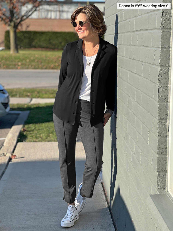Woman looking away while leaning against to a brick wall wearing Miik's Keeley notched collar long sleeve blouse in black opened, a white top and charcoal pants 