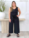 Miik model Christal (five feet three, size large) standing in front of a white wall smiling wearing Miik's Kimmay open-back capri jumpsuit in black